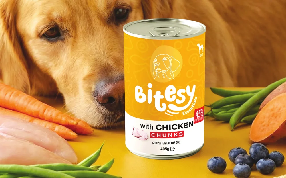 A golden retriever beside fresh meat and vegetables and a can of Bitesy Essentials